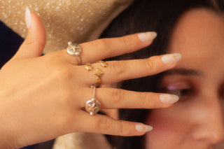 A close-up lifestyle shot of a person's hand wearing multiple rings, including a silver ring with a large star and a gold rings with 3 star details, against a dark background with a sparkly gold hat visible.