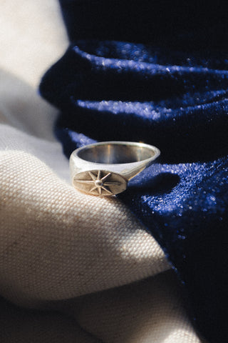 A silver signet ring with a starburst emblem on the face, resting on a textured cream linen fabric next to a deep navy blue velvet cloth.